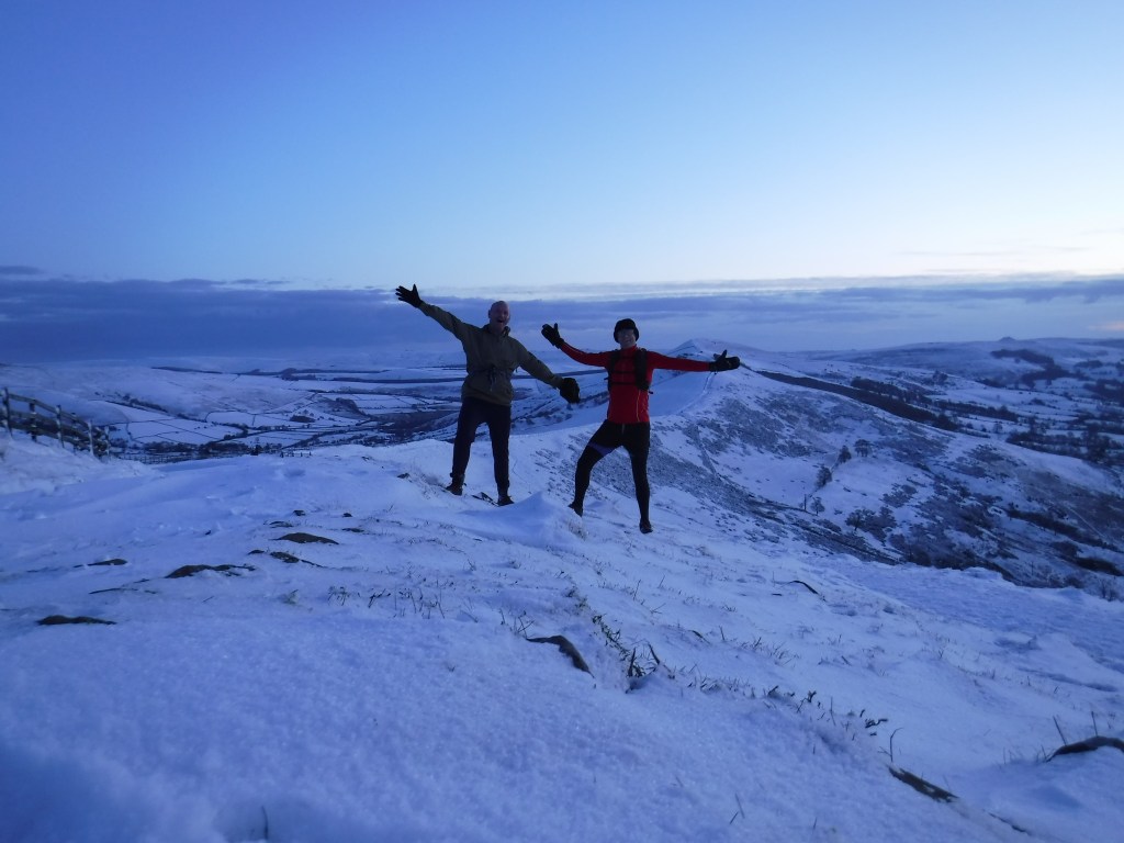 Two men, arms outstretched, stood on a snowy hill top around dawn.