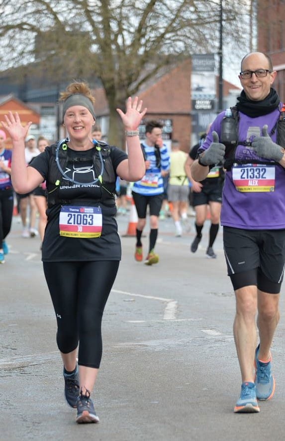 Jo Murray and Alex Masidlover on the course of Manchester Marathon. Jo is smiling and waving. Alex is smiling and holding thumbs up.